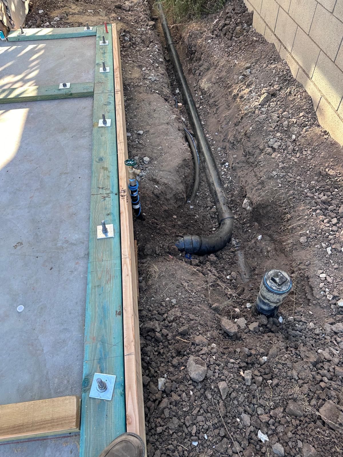 Open trench beside a new slab form, with a black drain pipe curving into the ground and a capped cleanout riser